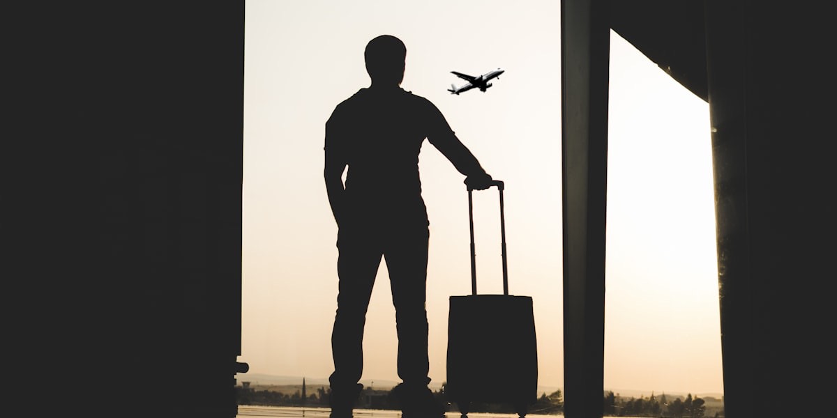 Silhouette d'un voyageur avec une valise devant une baie vitrée d'aéroport, un avion décollant au loin